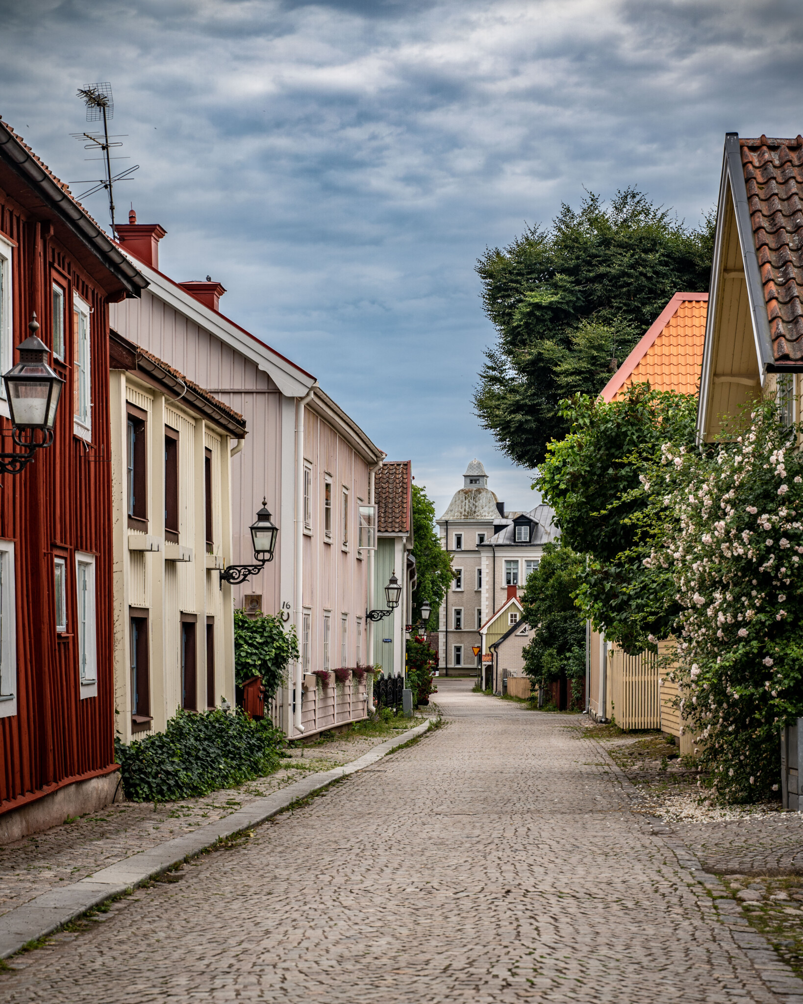 Kullertensgata med färgglada trähus i rosa, rött och gult i Vadstenas gamla stad, kantad av grönska och blommande buskar.