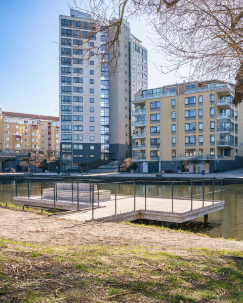 Träbrygga vid Stångån i centrala Linköping med moderna bostadshus i bakgrunden, en solig vårdag vid Stångebro