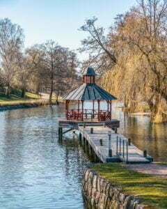 Röd lusthuspaviljong på brygga i Stångån, omgiven av hängpilar och grönska i centrala Linköping en tidig vårdag