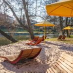 Solstolar i trä och gula parasoller på sandstrand vid Stångån i Linköping, omgivna av stora lövträd en solig vårdag.
