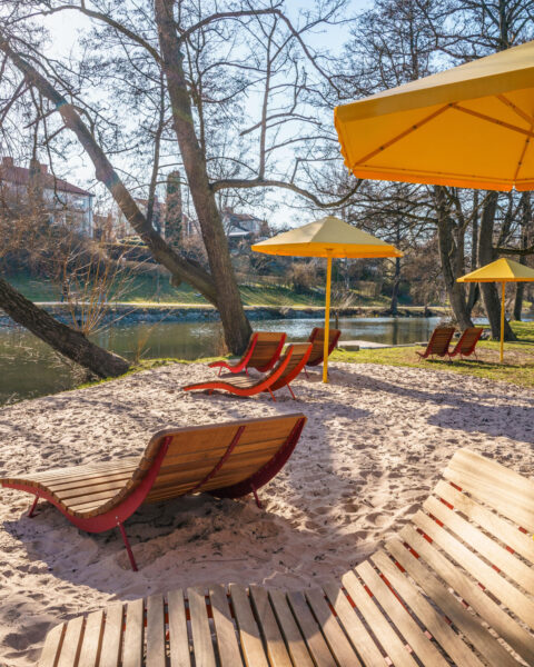 Solstolar i trä och gula parasoller på sandstrand vid Stångån i Linköping, omgivna av stora lövträd en solig vårdag.