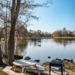 Picknicbord och grillplats vid Stångåns strand på Sågholmen i Linköping, med stilla vatten och kala vårträd i bakgrunden