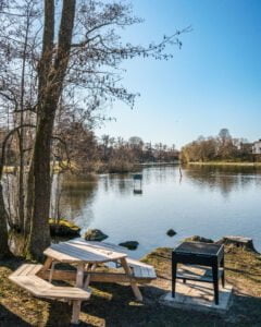 Picknicbord och grillplats vid Stångåns strand på Sågholmen i Linköping, med stilla vatten och kala vårträd i bakgrunden