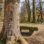 Trädskulptur med utskurna dörrar och fönster i en trädstam vid stenbänk i Grenadjärparken, Linköping, med damm i bakgrunden