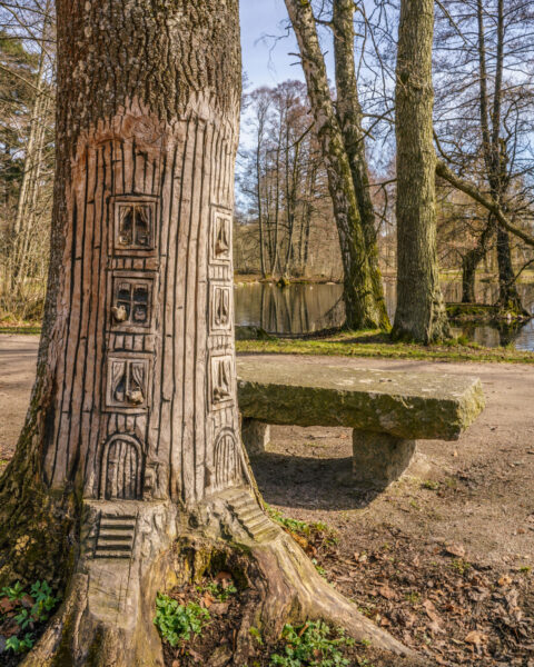 Trädskulptur med utskurna dörrar och fönster i en trädstam vid stenbänk i Grenadjärparken, Linköping, med damm i bakgrunden