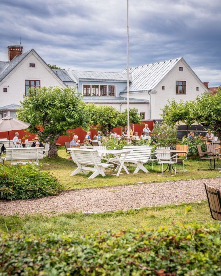 Gäster njuter av fika i den blomstrande trädgården vid Väderstad Centralkonditori med vita möbler, fruktträd och röda staket framför vita hus.