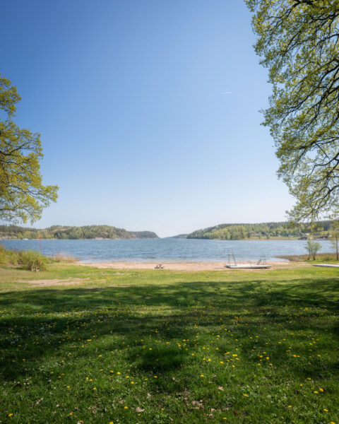 Grännäs havsbad i Valdemarsvik med grön gräsmatta, sandstrand, brygga och lugnt vatten omgivet av skogklädda höjder en solig vårdag