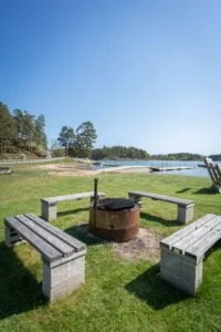 Grillplats med träbänkar på grönt gräs vid Sandgärdets havsbad i Gryt, med bryggor, sandstrand och skärgårdsvy i bakgrunden.