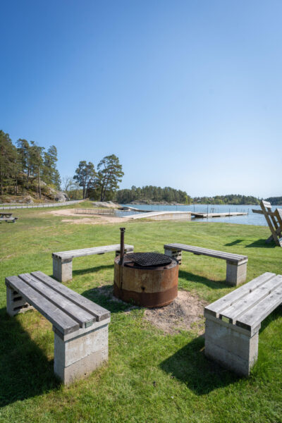 Grillplats med träbänkar på grönt gräs vid Sandgärdets havsbad i Gryt, med bryggor, sandstrand och skärgårdsvy i bakgrunden.