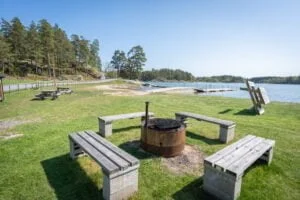 Grillplats med träbänkar på grön gräsmatta vid Sandgärdets havsbad i Gryt, med bryggor och skärgårdsvatten i bakgrunden.
