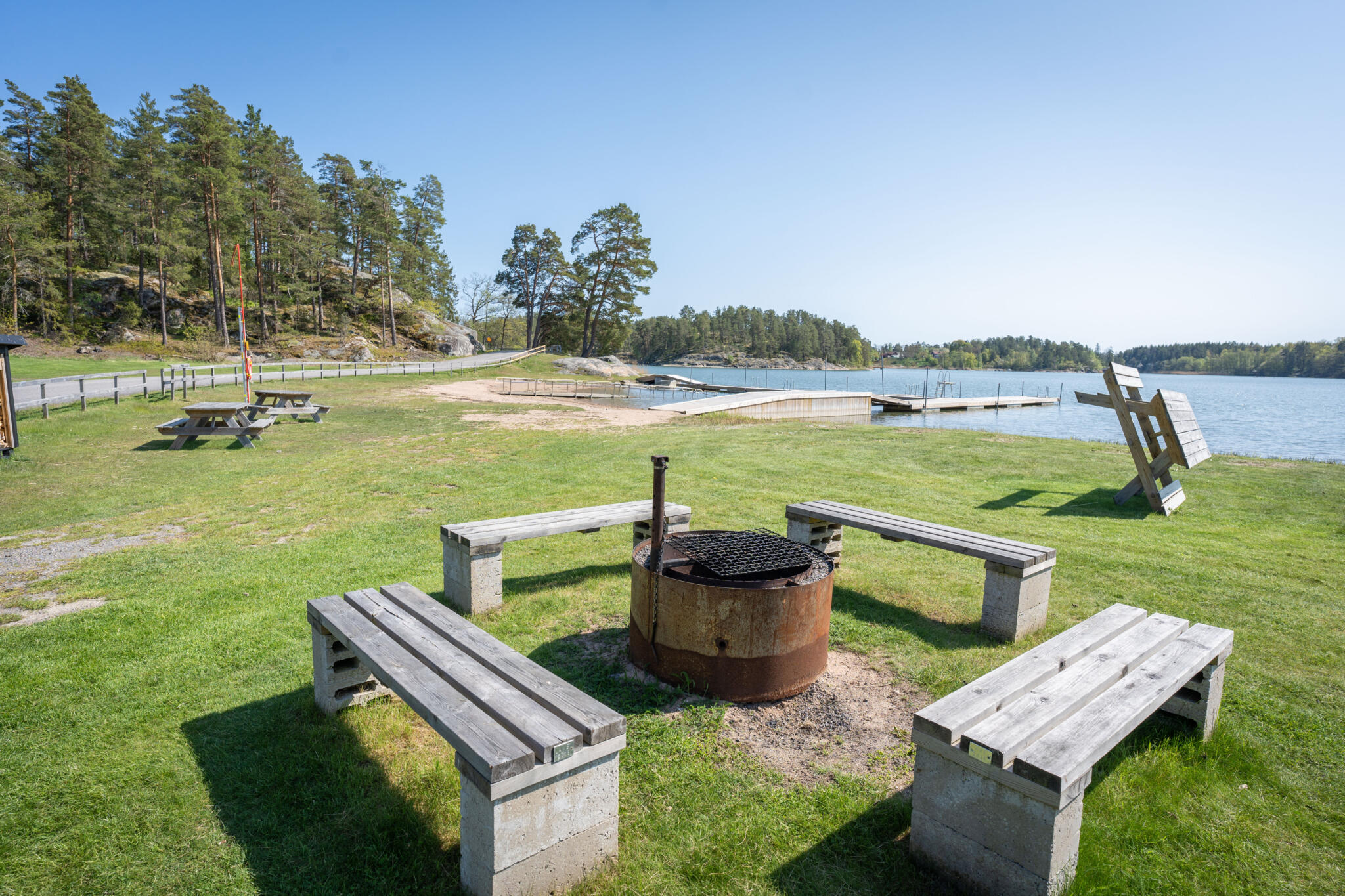 Grillplats med träbänkar på grön gräsmatta vid Sandgärdets havsbad i Gryt, med bryggor och skärgårdsvatten i bakgrunden.
