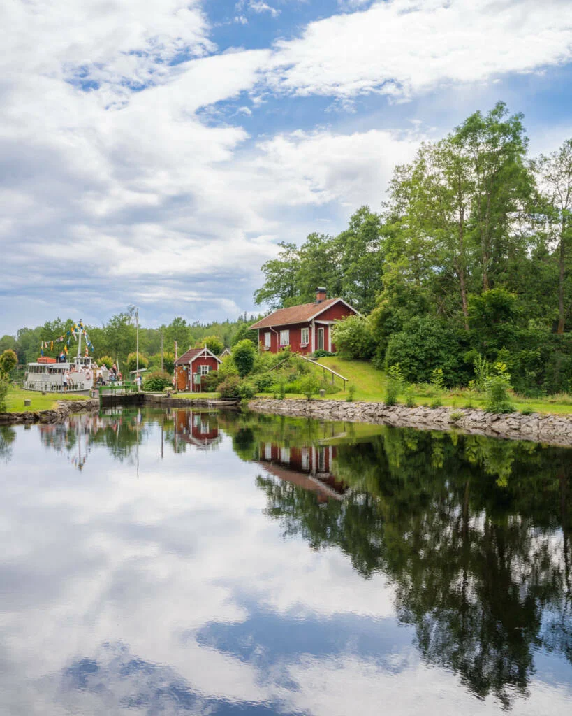 Röd slussvaktarstuga vid Dalslands kanal med spegelblank vattenyta, flaggprytt kanalbåt vid slussen och frodig grönska en sommardag.