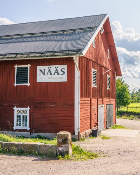 Röd ladugårdsbyggnad med skylt för Nääs Gårdsbryggeri i Ydre, omgiven av grönska en sommardag