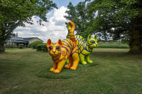 Två stora färgglada kattskulpturer i orange och grönt med blå mönster på en gräsmatta i Skulpturpark Vadstena, omgivna av lummiga träd