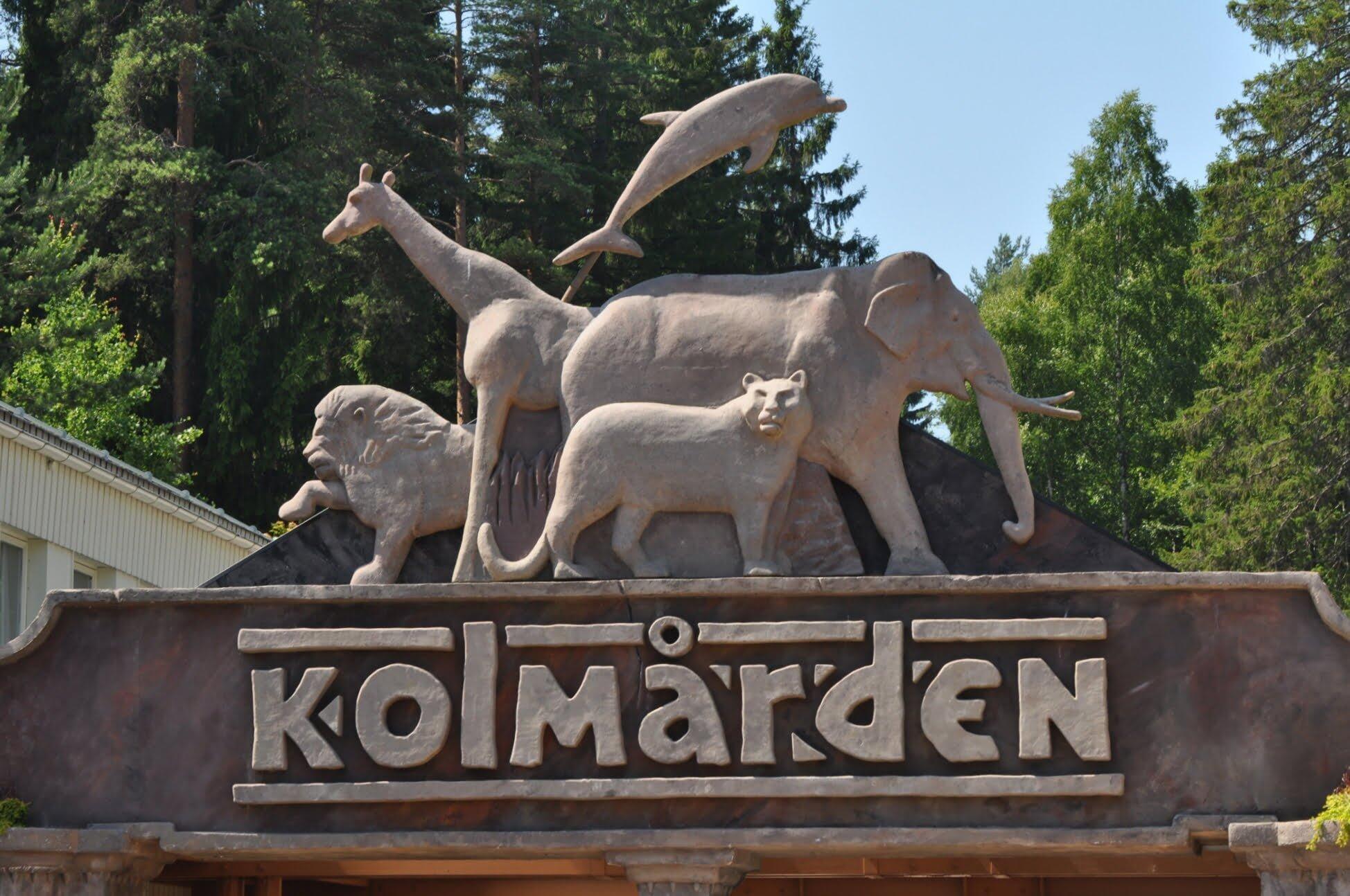 Entréskylt till Kolmårdens djurpark med skulpturer av lejon, giraff, delfin, elefant och leopard omgivna av grönska.