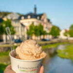 Hand som håller en bägare med italiensk glass från Konfekthörnan i Söderköping, med Storåns vatten och stadskärnan i bakgrunden.
