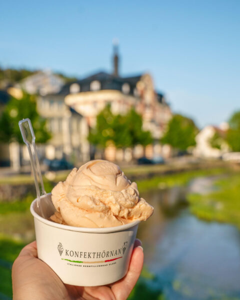 Hand som håller en bägare med italiensk glass från Konfekthörnan i Söderköping, med Storåns vatten och stadskärnan i bakgrunden.
