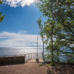 Utsiktsplats vid Stocklycke hamn på Omberg med solglittrande Vättern, dekorativt räcke och grönskande lövträd en sommardag