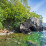 Klippformation med grottöppning vid Vätterns strand nära Stocklycke på Omberg, omgiven av frodig grönska och kristallklart vatten