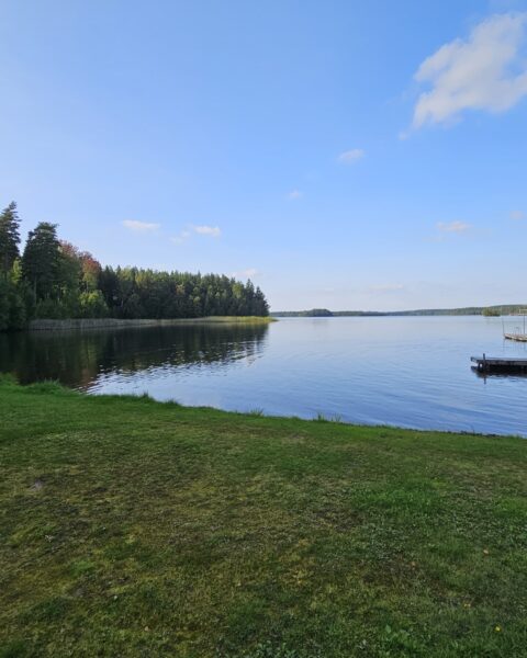 Lugn sjövy över Stora Sundsjön vid Sunds badplats i Ydre, med grön gräsmatta, brygga och skogsklädd strand under blå himmel.