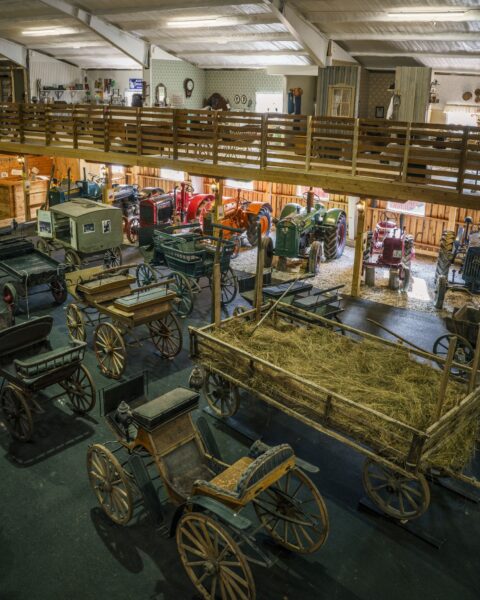 Lantbruksmuseet Ljusfallshammar med samling av gamla hästvagnar, hövagn och veterantraktorer i en rymlig lada med träräcke på övervåningen.