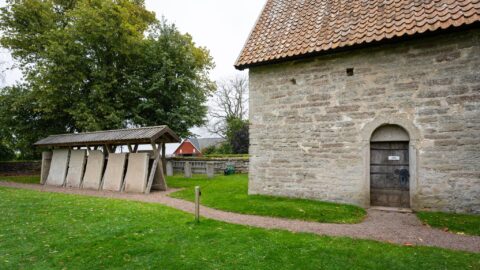 Örberga stenmuseum med medeltida stenhus och uppställda gravhällar under tak på grön kyrkogård, Vadstena kommun
