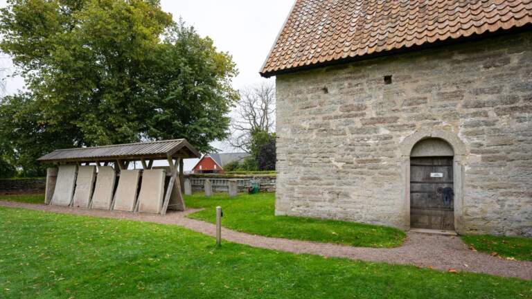 Örberga stenmuseum med medeltida stenhus och uppställda gravhällar under tak på grön kyrkogård, Vadstena kommun