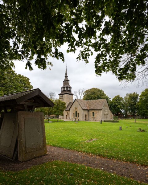 Örberga kyrka i Vadstena kommun med sitt medeltida stenkyrkoskepp och torn, sedd från kyrkogården med en gammal gravhäll under tak i förgrunden.