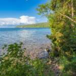 Klart vatten vid Älvarums udde på Omberg med stenig strand, frodig grönska och utsikt över Vättern en solig sommardag