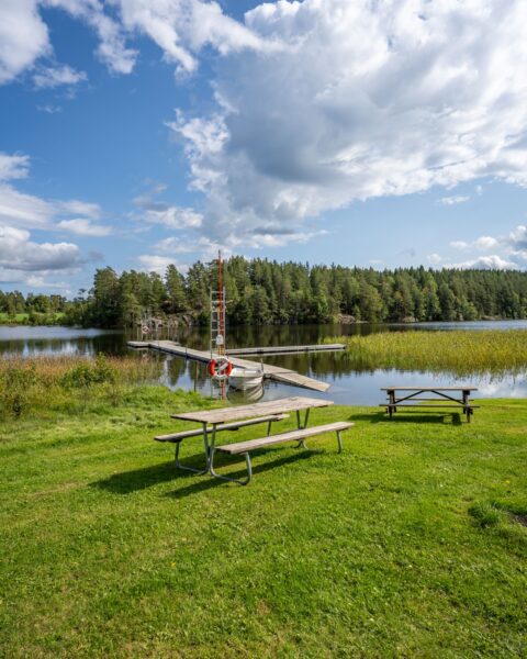 Näs badplats vid Raklången i Ydre med grön gräsmatta, picknickbord, badbrygga, livboj och skogsbeklädd strand en sommardag.
