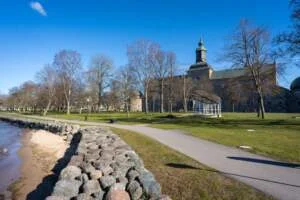 Strandpromenaden i Vadstena med stensatt strand, gröna gräsytor och avlövade träd, med Vadstena slott i bakgrunden en solig vårdag.