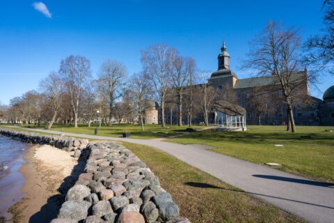 Strandpromenaden i Vadstena med stensatt strand, gröna gräsytor och avlövade träd, med Vadstena slott i bakgrunden en solig vårdag.