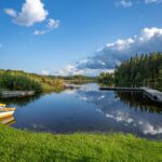 Stilla sjö vid Österby badplats i Ydre med brygga, roddbåtar, grön gräsmatta och barrskog som speglas i vattenytan en sommardag