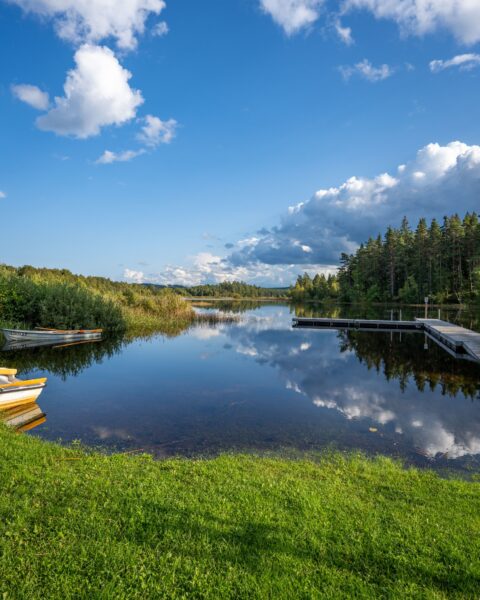 Stilla sjö vid Österby badplats i Ydre med brygga, roddbåtar, grön gräsmatta och barrskog som speglas i vattenytan en sommardag