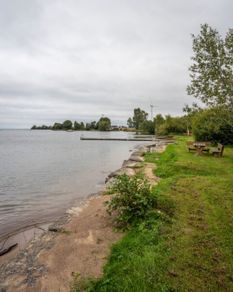 Strandkant vid Bårstad friluftsbad med sandstrand, gräsyta, sittbänk, brygga och utsikt över Vättern en mulen dag. Vindkraftverk i bakgrunden.