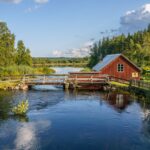 Röd smedja vid Österfors i Ydre, omgiven av strömmande vatten, träbro och grön norrländsk skog en sommarkväll