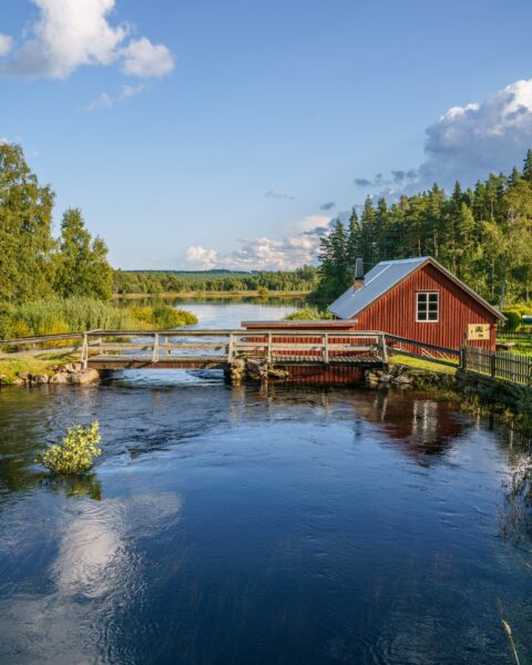 Röd smedja vid Österfors i Ydre, omgiven av strömmande vatten, träbro och grön norrländsk skog en sommarkväll