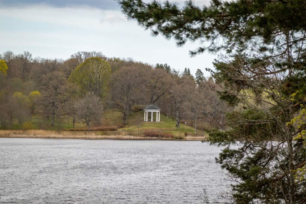 Klassiskt tempel med vita pelare vid Adelsnäs park i Åtvidaberg, omgivet av lövträd på en grön kulle vid sjön