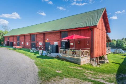 Stor röd ladugårdsbyggnad med grönt plåttak och träaltan med rött parasoll på Svarttorps Gård vid sjön Hunn i Finspång, en solig sommardag.
