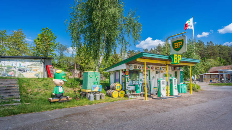 Nostalgisk BP-mack i Björkfors från 1950-60-talet med gröna bensinpumpar, BP-skylt, maskot och lummig grönska en solig dag.