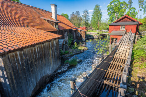 Strömmande vatten mellan historiska bruksbyggnader vid Häfla hammarsmedja, med vattenhjul, träränna och röda hus en solig sommardag i Östergötland