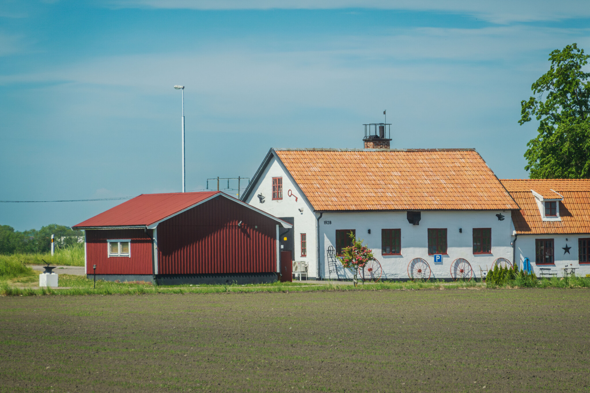 Hagdahls smide vid öppet åkerlandskap – vit byggnad med tegeltak och dekorativa vagnshjul på fasaden, intill ett rött uthus.