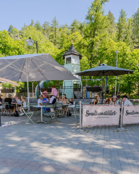 Gäster njuter av glass på uteserveringen vid Smultronstället Glassrestaurang i Söderköping, med parasoller och grönska en solig sommardag.