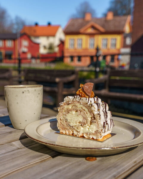 En krämig Budapestbakelse serveras vid Svartån i Mjölby, med färgglada trähus i bakgrunden.