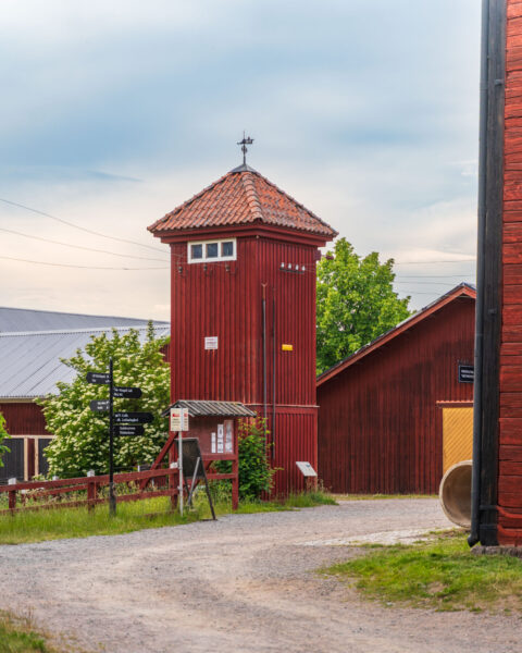 Rödfärgad träbyggnad med torn och vindflöjel vid Valla Gård i Linköping, omgiven av grusväg och grönskande träd.