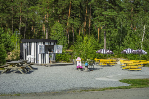 Svartvitrandig glasskiosk med gula picknickbord och randiga parasoll på grusgård omgiven av tallskog vid Sänkdalens Gård.