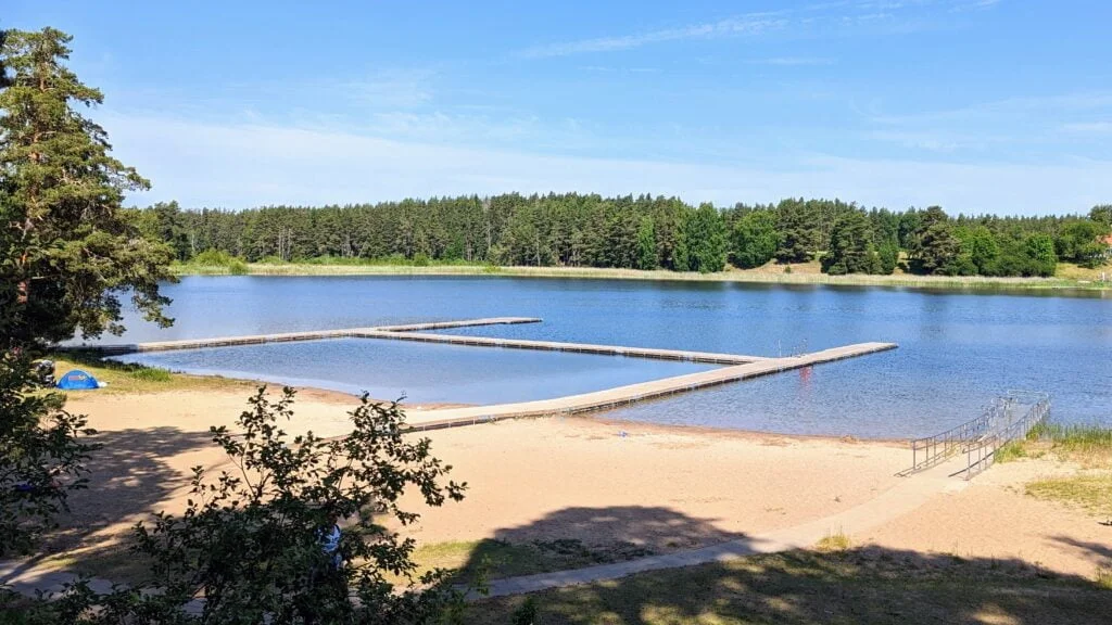 Sandstrand vid Skogssjöns badplats i Mjölby med bryggor som bildar en bassäng i det stilla vattnet, omgivet av tallskog en solig sommardag.