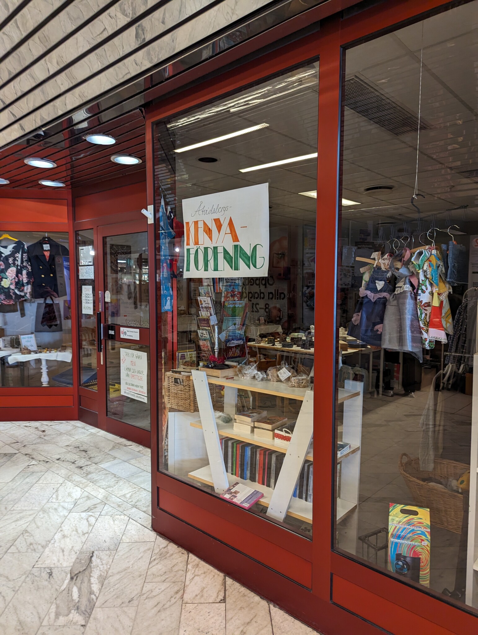 Skyltfönster med röda karmar till Åtvidabergs Kenyaförening i Getinggallerian, med kläder, böcker och hantverk i fönstret.