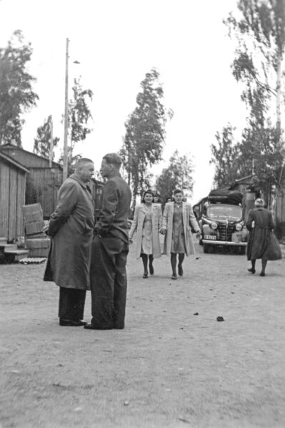 Svartvitt foto från flyktingförläggningen i Doverstorp 1945 med personer på en grusväg, björkar, träbaracker och en parkerad bil från 1940-talet.