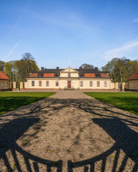 Lambohovs säteri i Linköping, ett gulputsat lustslott i rokokostil med grusgång, gröna gräsmattor och flygelbyggnader under klarblå himmel.
